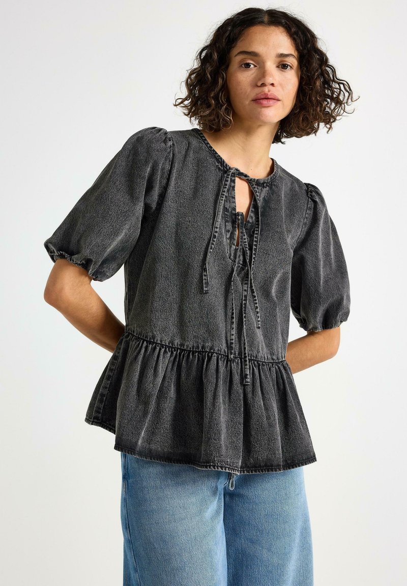 Kvinde iført en falmet sort bluse med puffærmer og peplum-kant samt snøredetaljer, parret med lyseblå jeans, stående foran en hvid baggrund.