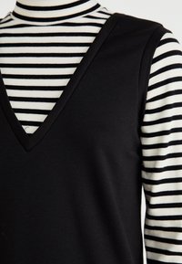 Vestido negro sin mangas con un profundo escote en V, superpuesto a una camisa de rayas blanca y negra de manga larga. Textura suave con patrones contrastantes.