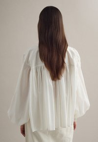 Persona con cabello castaño largo que lleva una blusa blanca plisada y suelta, de pie de espaldas sobre un fondo beige liso.