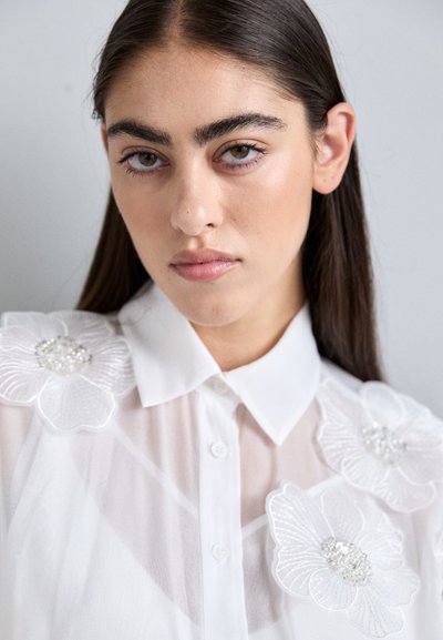 Hvid gennemsigtig bluse med krave, der har blomsterbroderi med perleaccent på skuldrene. Glat tekstur og let design.