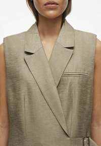 Chaleco sin mangas beige con un acabado texturizado, collar ancho y un bolsillo único. Presenta un corte entallado y un diseño minimalista.