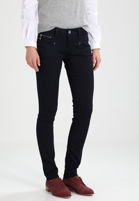 Jeans azul oscuro de mezclilla, con un corte ajustado, bolsillo con cremallera y detalles de botón sutiles. Combinados con un chaleco gris y zapatos rojos.