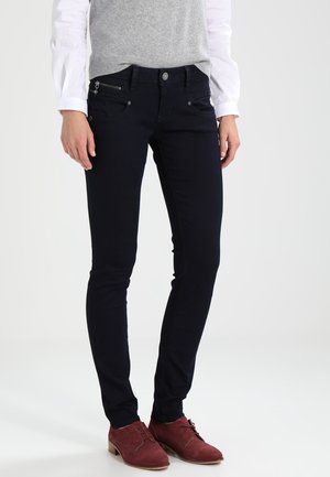 Jean en denim bleu foncé, avec une coupe slim, une poche zippée et des détails de bouton discrets. Porté avec un gilet gris et des chaussures rouges.