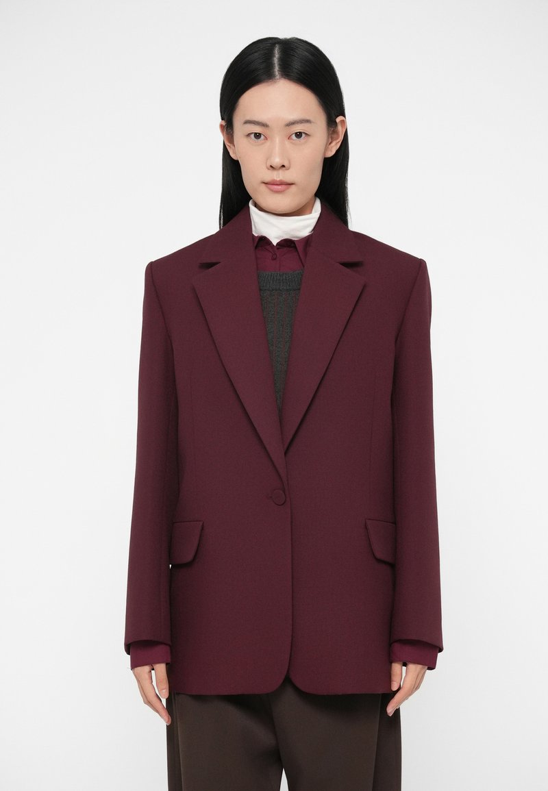 Blazer su misura bordeaux con rever a punta, due tasche frontali, spalle strutturate e chiusura con bottoni. Indossato sopra un outfit stratificato grigio e bianco.