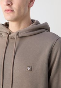Sudadera marrón con acabado texturizado, que cuenta con un cordón y un logotipo cuadrado bordado en el pecho.