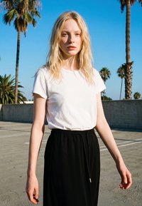 Jauna sieviete ar gariem blondiem matiem, valkājot baltu T-kreklu un melnu svārku, stāv ārā ar palmām un zilu debesu fonu aiz muguras.