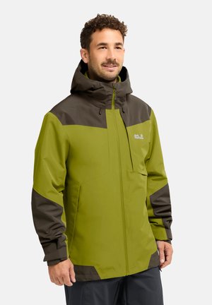 Mann mit kurzen lockigen Haaren, der eine grüne und dunkelbraune Kapuzenjacke mit Frontreißverschluss und kleinem Logo auf der Brusttasche trägt.