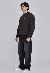 Brązowa bluza z okrągłym dekoltem i haftowanym logo, w zestawieniu z ciemnymi dżinsami i beżowymi sneakersami. Model stoi na jednolitym tle.