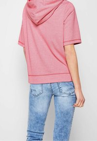 Person iført lyseblå denim jeans og en løs pink kortærmet hættebluse, stående med ryggen imod kameraet.