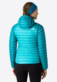 Turkos isolerad jacka med quiltat mönster, en åtsittande huva och axellogo. Slät textur med kontrasterande sömdetaljer.