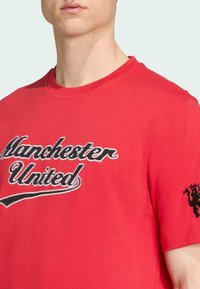 Camiseta de algodón roja con la frase "Manchester United" en texto bordado en blanco y negro, y un logo negro en la manga derecha.