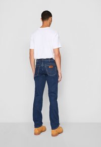 Donkerblauwe denim jeans met een rechte snit, zichtbare stiksels en een leren label op de tailleband, gecombineerd met een eenvoudig wit T-shirt en lichtgekleurde laarzen.