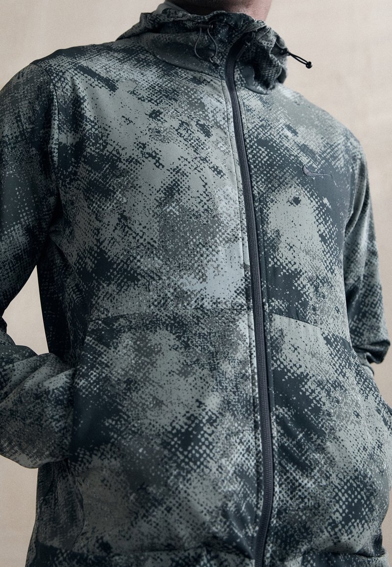 Veste à capuche avec un motif pixellisé gris et noir, fabriquée en matériau léger, dotée d'une fermeture éclair à l'avant et de poches latérales.
