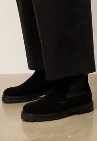 Fekete szarvasbőr Chelsea csizmák vastag gumitalppal, fekete, cropped nadrággal viselve, fényes padlón állva.