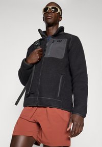 Schwarze Fleecejacke mit grauen Akzenten, mit Reißverschlusstaschen und hohem Kragen, kombiniert mit roten Shorts. Sonnenbrille und Schultergurt sichtbar.