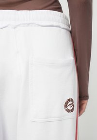 White sweatpants with an elastic waistband, featuring a patch pocket and a brown logo detail. Smooth texture and flat seams. Red side stripe.