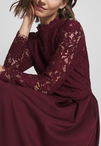 Top en dentelle bordeaux à manches longues, avec des motifs floraux et un col en dentelle ondulé, associé à une jupe fluide bordeaux.