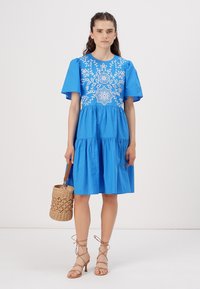 Blue cotton dress with short puff sleeves, white embroidery on the bodice, tiered skirt, and a textured wicker handbag. Beige strappy heels.