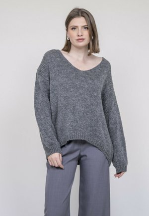 Femme aux cheveux raides mi-longs portant un pull gris ample et un pantalon gris à jambes larges, debout devant un fond gris uni.