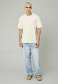 T-shirt beige à manches courtes avec un petit graphisme sur la poitrine gauche, associé à un jean baggy bleu clair et des chaussures beiges.