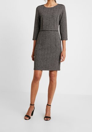 Vrouw draagt een grijze, knielange, nauwsluitende jurk met textuur, lange mouwen en zwarte hoge hakken met enkelbandje tegen een witte achtergrond.