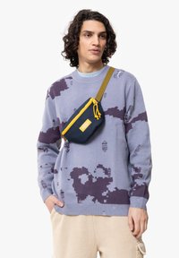 Maglione lilla con motivi astratti viola scuro e dettagli consumati, abbinato a una borsa a tracolla blu navy con accenti gialli.