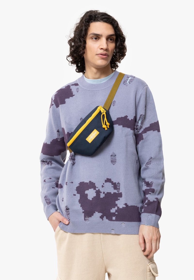 Maglione lilla con motivi astratti viola scuro e dettagli consumati, abbinato a una borsa a tracolla blu navy con accenti gialli.