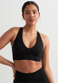 Svart sports-BH med dyp V-utringning, sømløst design og brede stropper, laget av et glatt stoff. Har en tettsittende silhuett.