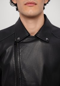 Jeune personne portant une veste en cuir noire avec un col à boutons-pression et une fermeture éclair à l'avant, montrant le haut de la poitrine et la zone du cou.