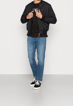 Man wearing black bomber jacket over black graphic t-shirt, blue jeans, and black-and-white sneakers standing against a white background.