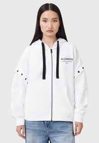 Sudadera blanca con cremallera y logo negro, cordones y detalles a presión en las mangas. Material suave, corte relajado y bolsillos frontales.