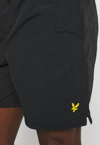 Pantaloni corti neri realizzati in tessuto leggero e testurizzato, con un ricamo decorativo di un'aquila gialla sul lato sinistro in basso.
