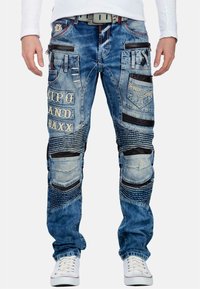 Jeans in denim blu strappati con texture varie, toppe nere, logo ricamato e molteplici tasche per dettagli e funzionalità.