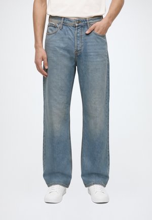 Personne portant un jean large bleu clair, des baskets blanches et une chemise blanche, une main dans la poche, debout devant un fond uni.