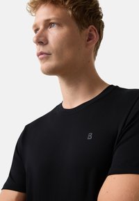 Młody mężczyzna o jasnej karnacji i krótkich blond włosach, ubrany w prostą czarną koszulkę z okrągłym dekoltem, z małym szarym logo "B" na piersi.