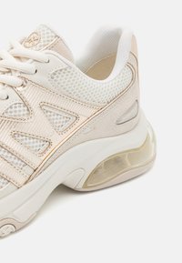 Baskets beige et blanches avec des détails en mesh et en daim, semelle épaisse et rembourrée, avec une poche d’air transparente au niveau du talon.