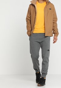 Brun vattentålig jacka, gul t-shirt, grå sweatpants och svarta sneakers. Har logotyp på bröstet och byxorna, elastiska mudd och midja.