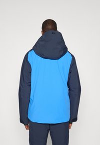 Kék és sötétkék vízálló dzseki kapucnival, sima textúrával és kontrasztos színblokkokkal; logó a hátulján.