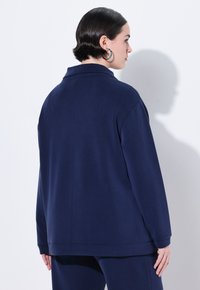 Maglione blu navy in tessuto liscio, con colletto, spalle scese e vestibilità comoda con orlo a fascetta. La vista posteriore mostra dettagli di design semplici.