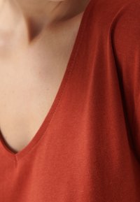 Rdzawoczerwony top z dekoltem w serek o gładkiej bawełnianej teksturze i widocznym przeszyciu wzdłuż dekoltu, prezentujący wygodne dopasowanie.