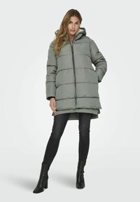 Padded green coat with a hood, featuring a front zipper, quilted design, and side pockets; paired with black skinny jeans and heeled boots.