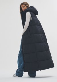 Fekete, párnázott, hosszú mellény kapucnival, steppelt textúrával és elöl cipzárral. Enyhén hosszú ujjú felső és bő kék farmer felett viselve.