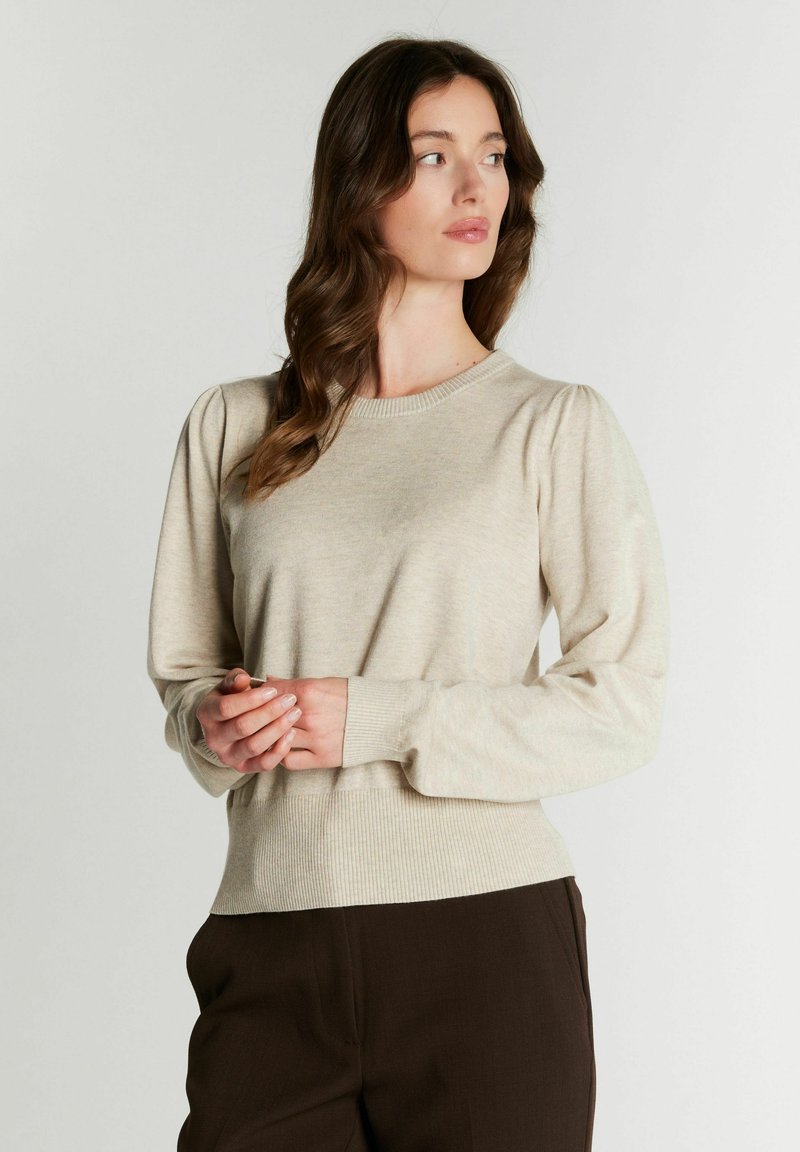 Femme aux longs cheveux bruns portant un pull beige à manches longues et un pantalon foncé, regardant sur le côté avec les mains jointes.