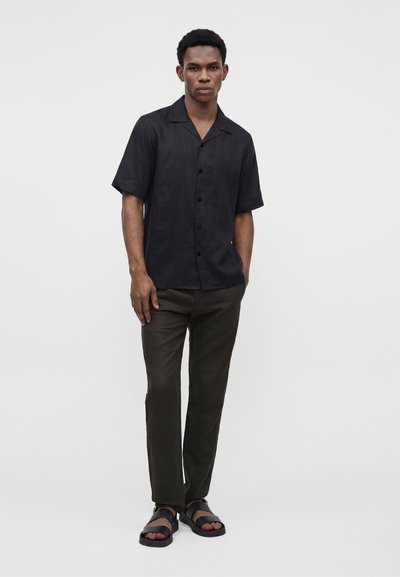 Jeune homme debout, les mains dans les poches, portant une chemise noire à manches courtes boutonnée, un pantalon foncé et des sandales noires sur un fond uni.
