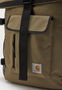 Mochila de tela color caqui con bolsillo frontal, cremallera negra, correa negra y parche blanco con logo y emblema naranja con el texto "carhartt".