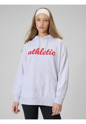 Młoda kobieta ubrana w jasno szarą bluzę z kapturem z czerwonym napisem "athletic", czarne legginsy, białą opaskę na głowę i srebrne kolczyki koła na szarym tle.
