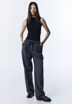 Donna con capelli lunghi e lisci che indossa un top nero senza maniche, pantaloni grigi con coulisse e mocassini neri con dettagli decorativi.