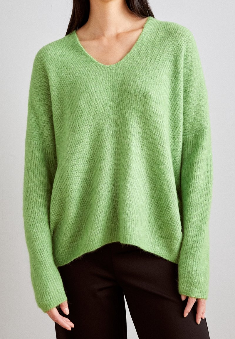 Femme portant un pull vert clair à col en V, ample et à texture douce, avec des manches longues et un pantalon noir, debout devant un fond uni.