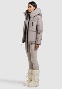 Lichtgrijze pufferjack met een hoge kraag, ritszakken en horizontale stiksels. Gecombineerd met beige leggings en witte sneeuwlaarzen.