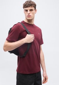 Νεαρός άνδρας με βουργουνδί μπλούζα και μαύρο σακίδιο πλάτης, κρατώντας ένα λουρί στον ώμο του, σε απλό φωτεινό φόντο.